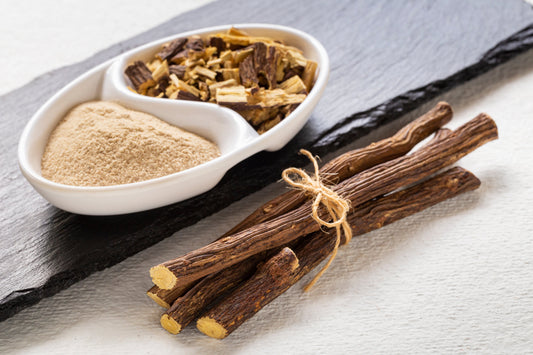 Dried licorice root sticks tied with twine, alongside a white dish containing powdered licorice and chopped licorice root pieces, displayed on a slate board.